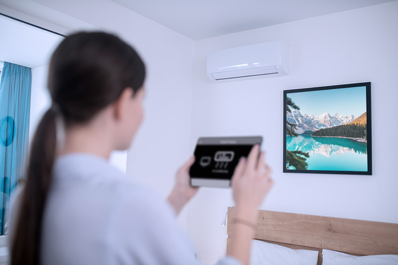 Back view of a dark-haired lady with the electronic device in the hands standing before the wall-mounted air conditioning unit