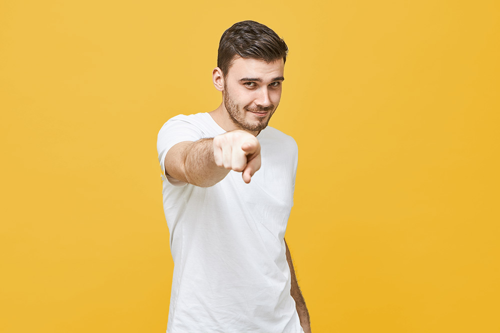 Picture of attractive self confident young brunette male in white casual t-shirt looking straight at camera and pointing fore finger, giving you confidence, having brilliant idea. Selective focus